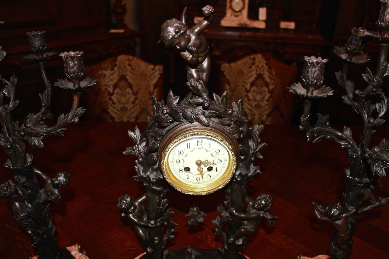This three piece French clock set is made from spelter and marble.  The set includes a pair of candelabras along with the main clock.  Each piece is made in an ornate style that depicts figural and organic details.  The clock features an exposed