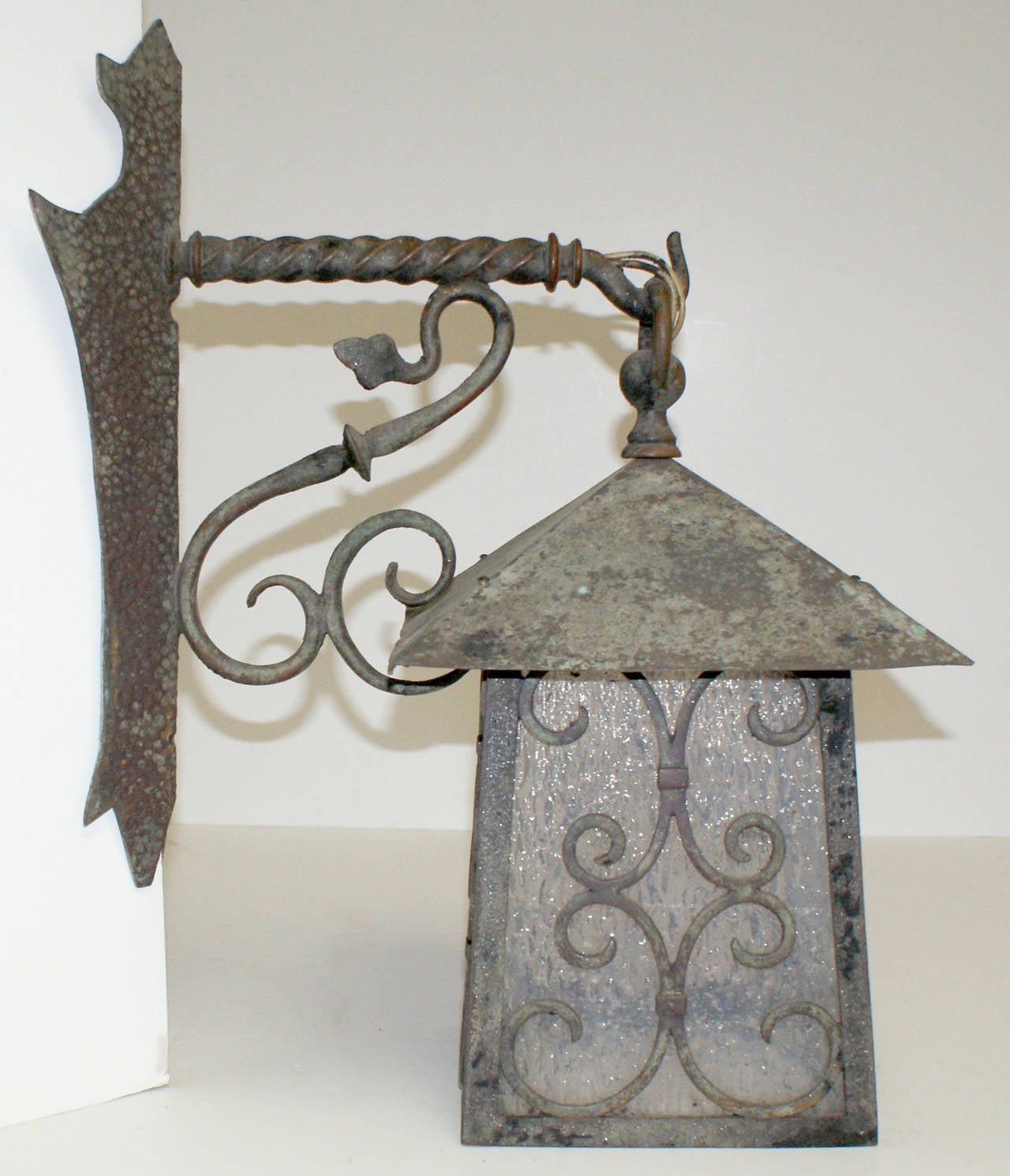 Nicely patinated antique bronze Arts and Crafts style corner mount exterior lantern sconce having hammered backplate, decorative scrolled metalwork and textured glass shade.