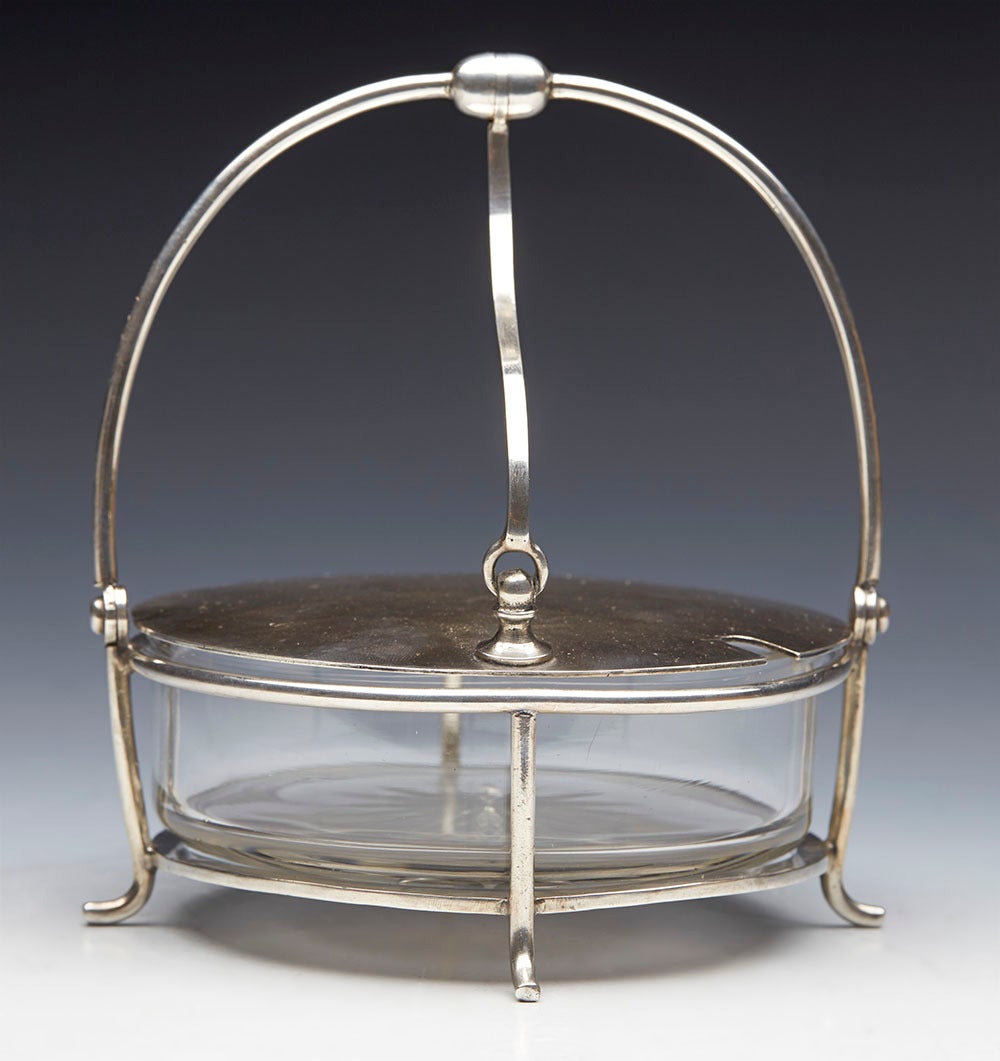 We offer this rare and stylish Arts and Crafts preserve pot designed by Christopher Dresser with a glass dish set within a silver plated frame and dating from around 1890. The preserve pot stands raised on four legs with a rounded lift out glass