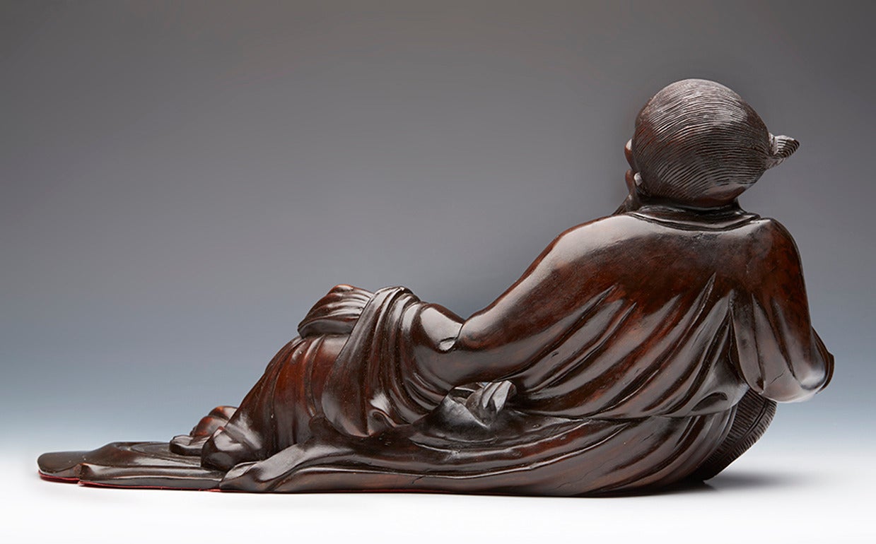 An antique lacquered hard wood figure of a reclining immortal. He is resting on a basket and with a miniature fisherman presenting him a large fish. The figures are disproportionate to each other and the carver possibly is reflecting on the