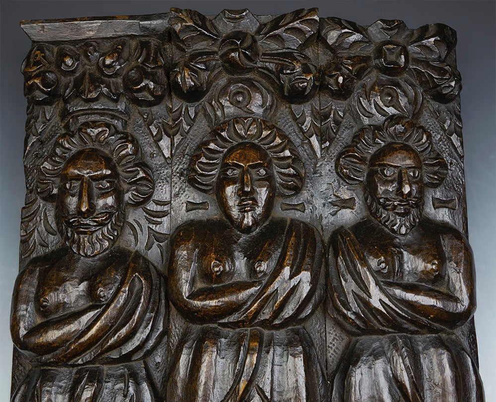 A carved architectural wooden panel mounted on a pedestal base dating from the 17th century. The hard wood panel is carved in deep relief from three pieces of wood. The three figures could possibly be saints, stood beneath floral garlands. The panel