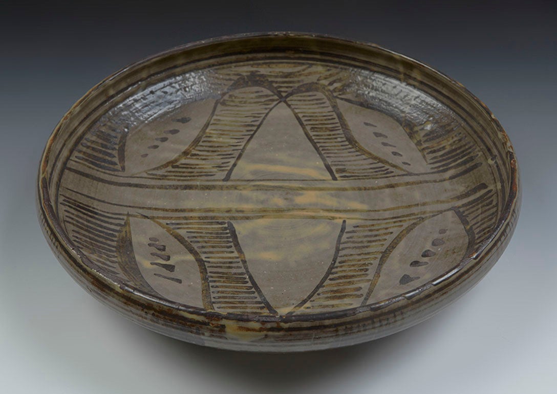 A bowl by Michael Cardew during his time at the Abuja Pottery in Nigeria where Cardew taught for a time. His work from this period often incorporates African style designs. This stoneware bowl is glazed in a brown slip with a leaf and stripe design.