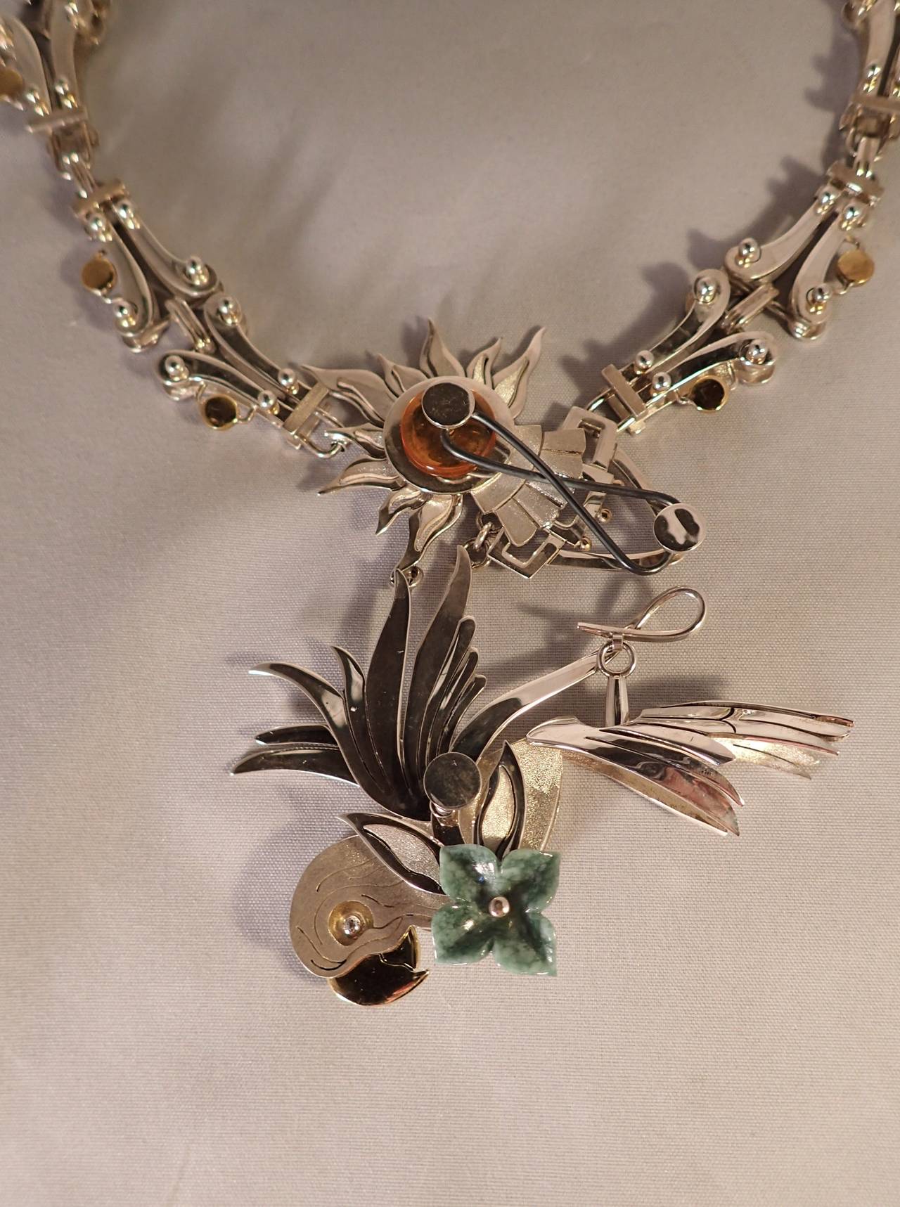 Parrot I by Sergio Gomez 
Necklace
925 Silver, gold, carved jade and amber, feather.
A magnificent sculptural silver and gold parrot in flight pendent carrying a wonderfully carved jade flower, suspended from a fantastic sun and heavens