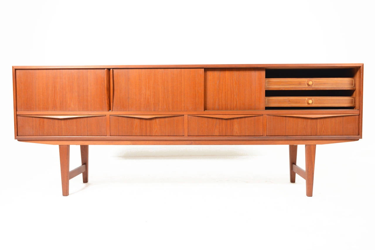 This low Danish modern mid century credenza crafted in teak was designed by E.W. Bach for Sejling Skabe. Left and center doors slide open to reveal open storage while the right interior features two removable drawers. Four drawers with dipped teak
