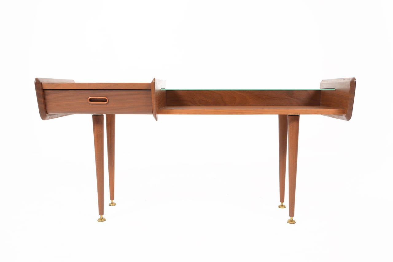 This Belgian modern entryway console table is constructed in teak and features a drawer and removable glass shelf. The simple yet elegant design is perfect for any modern entry, foyer, or living room. Legs are crafted from turned teak and wear their