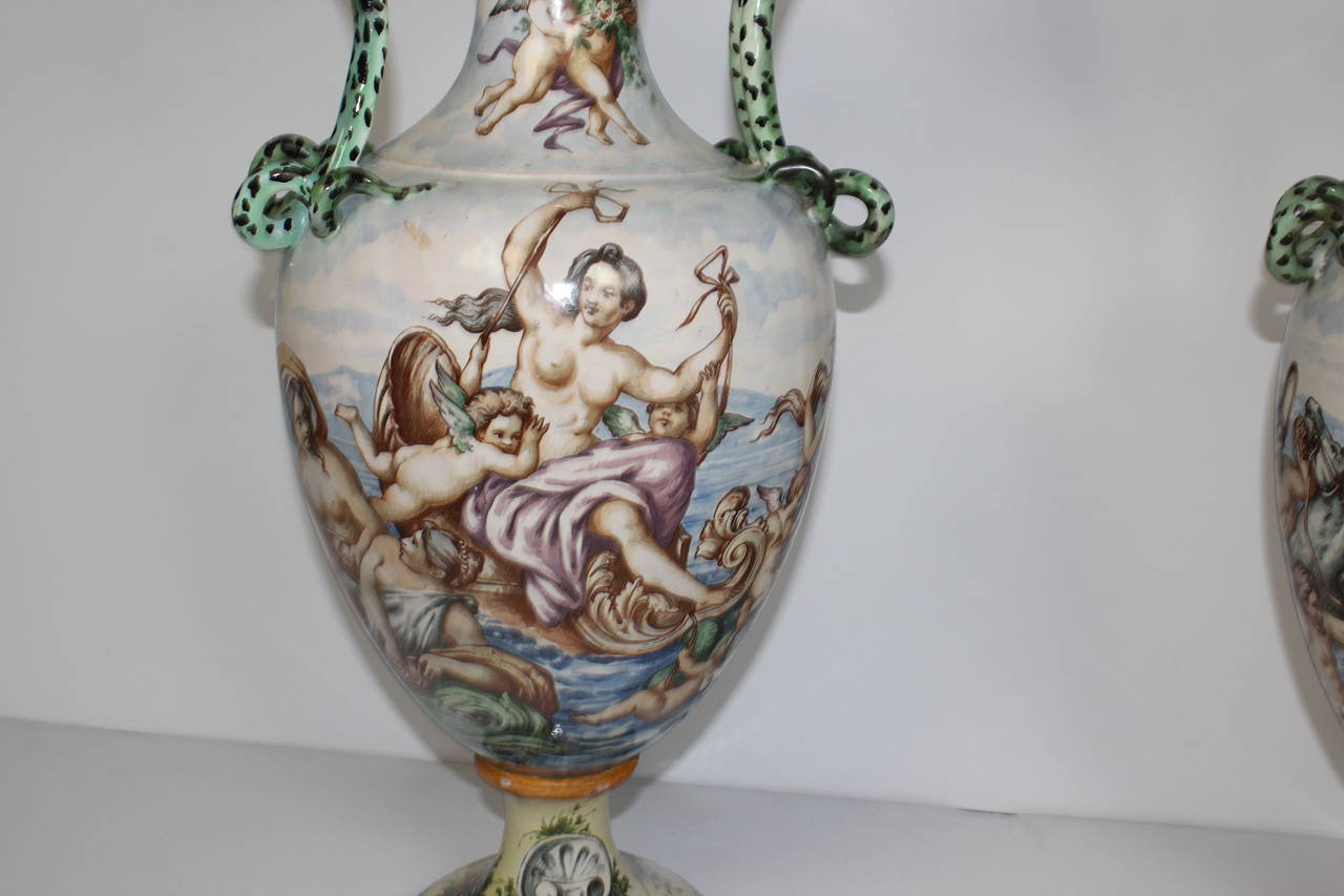 Late 19th century hand-painted Italian urns. The urns depict men, women, and angles in the ocean. The colors are soft but the handlers are painted in green and black in snack pattern. They are both signed.