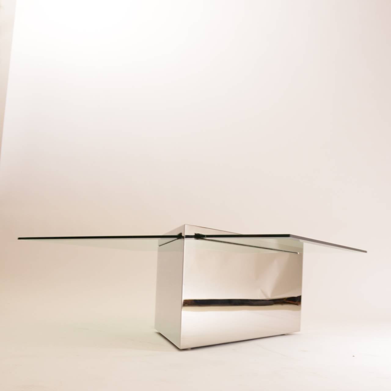 A cantilevered low glass table supported by a satin stainless steel seamless monolithic base with pencil edges. The solid mass of the top is intersected by a 1/2