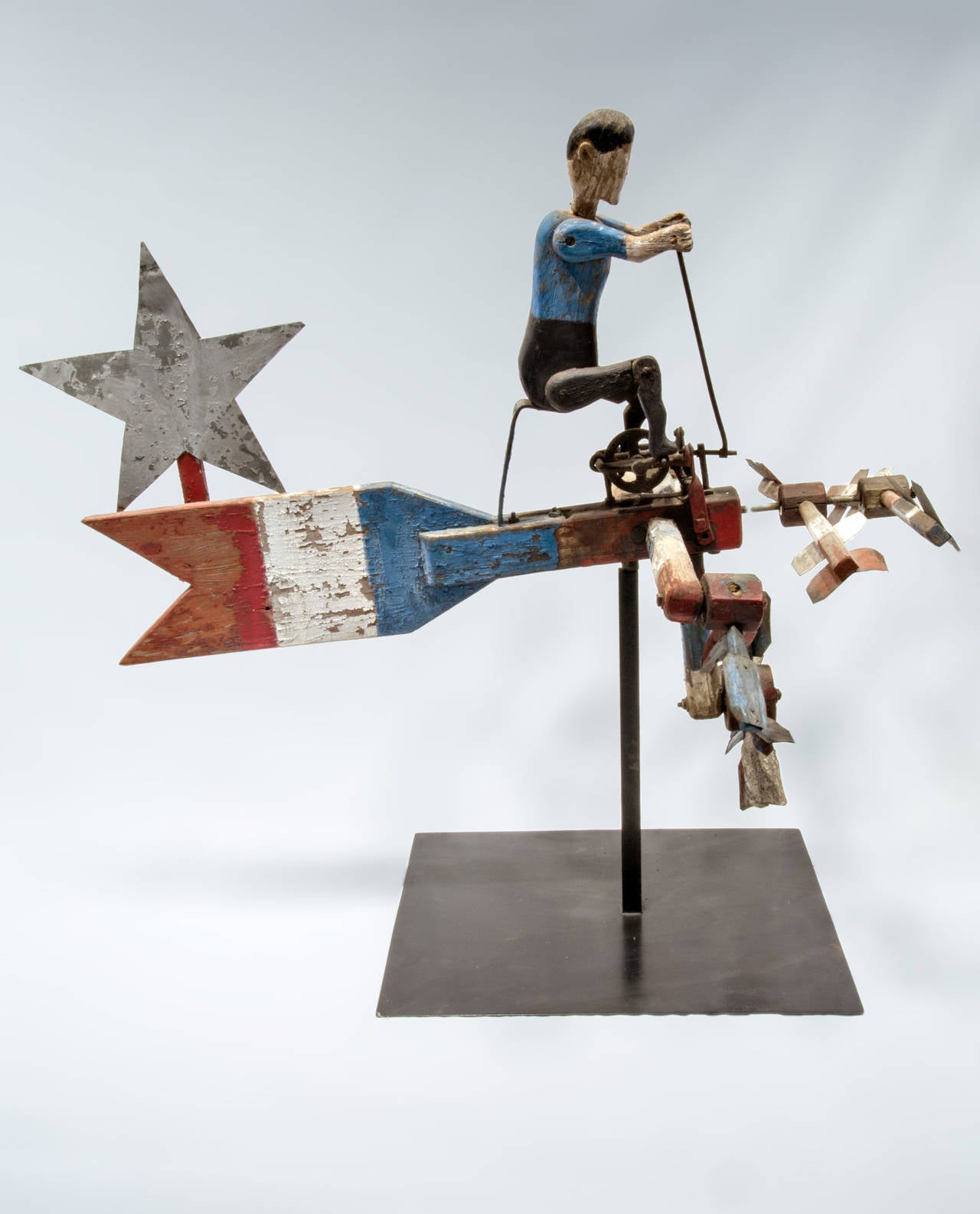 Carved pine whirligig of man on a bicycle, propeller driven and painted in patriotic colors with painted sheet metal star attached to arrow. The carved figure has articulated arms and legs and is seated on a metal stand. All original with minor