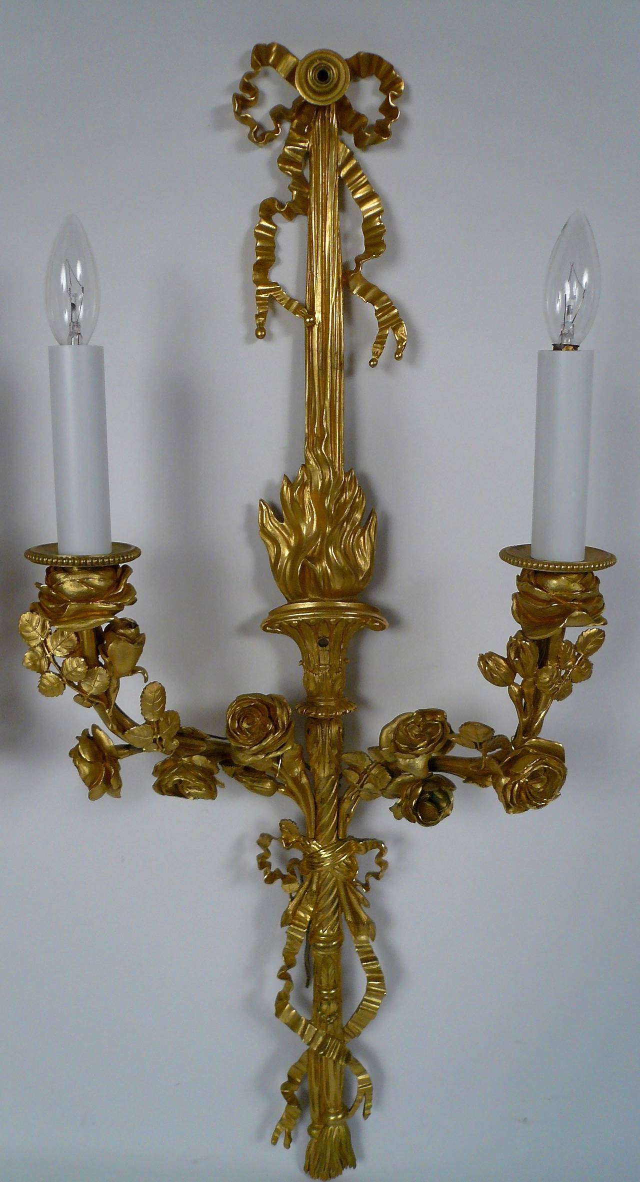 Pair of E. F. Caldwell gilt bronze Louis XVI style two-light sconces of bow knot and torch motif, with foliate arms terminating in rose blossom candle cups.