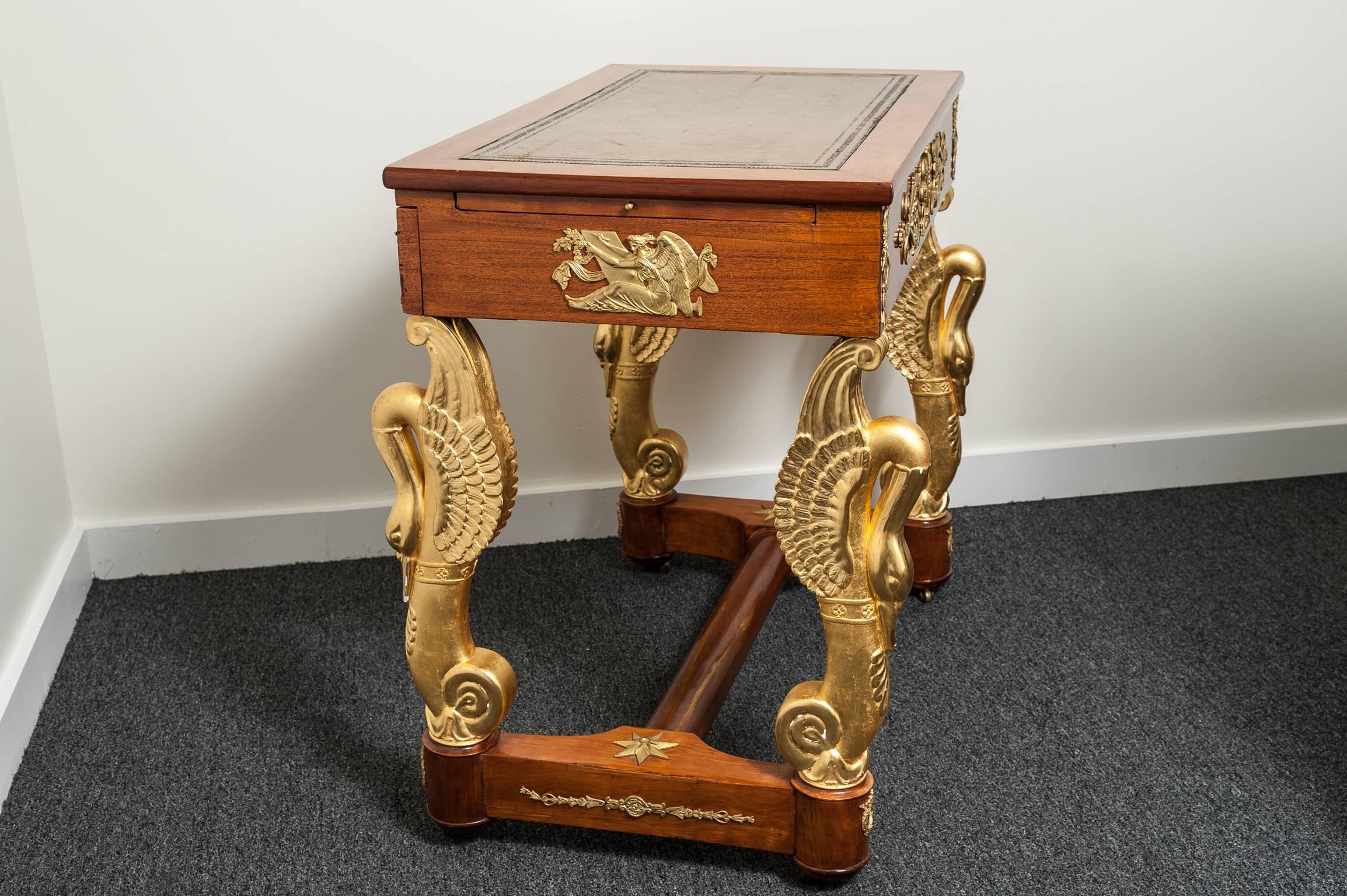 This stylish and chic French Empire piece dates from the napoleioic period of the early 19th century and is fabricated in mahogany with inset leather top, elongated, hand-carved swans with gold-gilt and bronze dore mounts depicting classical scenes