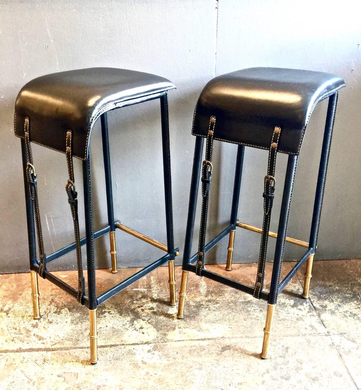 This is a superb pair of sexy Jacques Adnet bar stools that date to the 1950s. The Fine leather seats are strapped to the foot rails with horse tack buckles that have just the right amount of patina. The hand stitching is in great condition as are
