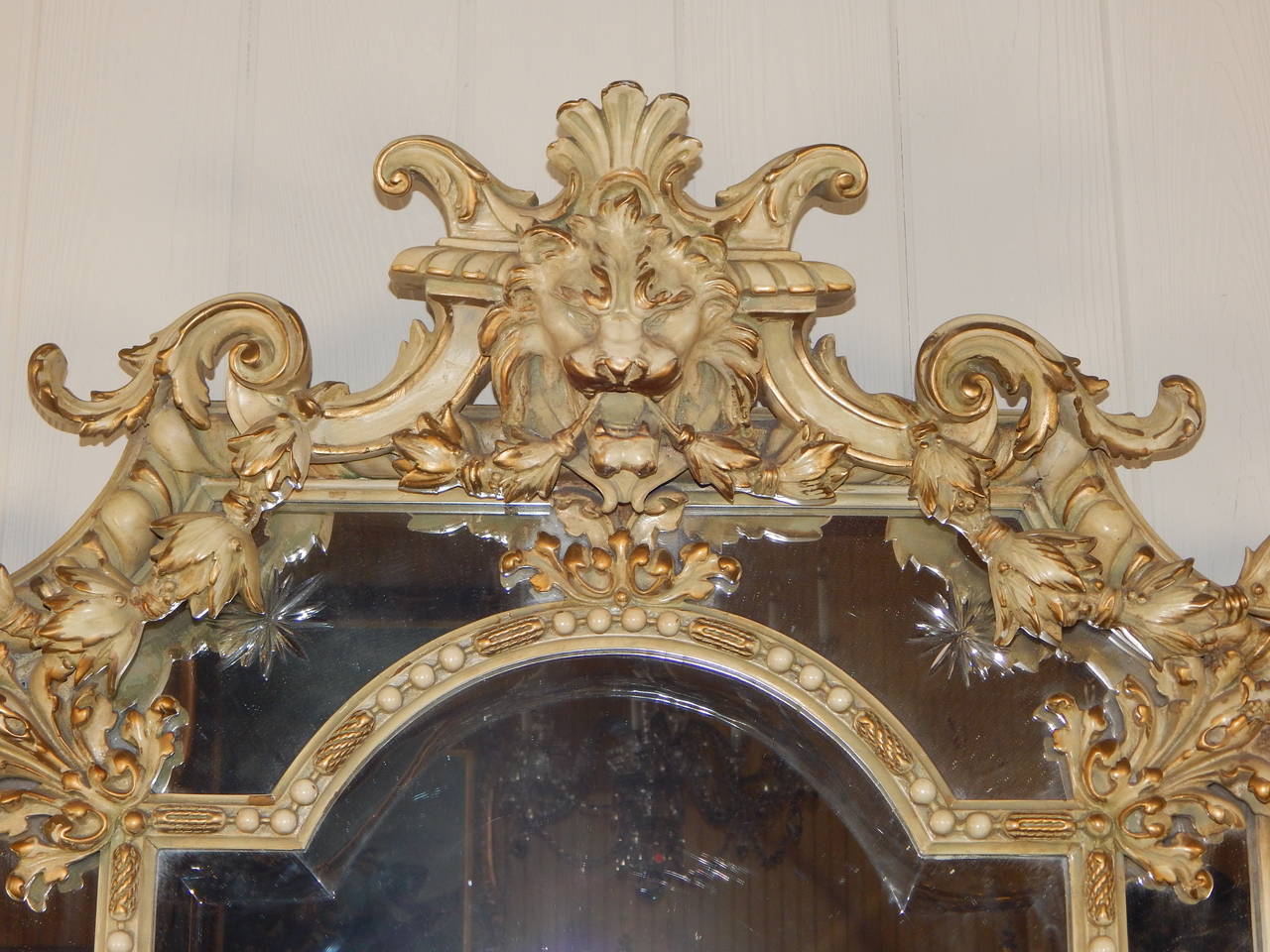 Fantastic cream colored enamel painted carved mirror with gilt highlights, and an incredible lion's face at the top, with a border of scrolls and ornaments. It also features beveled glass, along with incised cut glass stars.