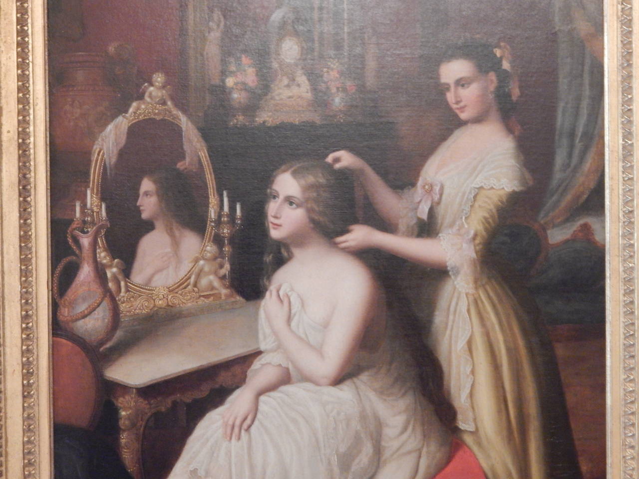 A beautifully executed oil on canvas of a young woman getting help with her hair, while sitting at a dressing table. Unsigned.