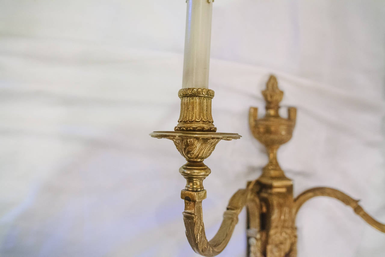 This incredible pair of Louis XVI style French bronze two-arm sconces features an urn finial at the top with intricate foliate details on the arms attaching to a tapering plinth. These sconces have been newly rewired.