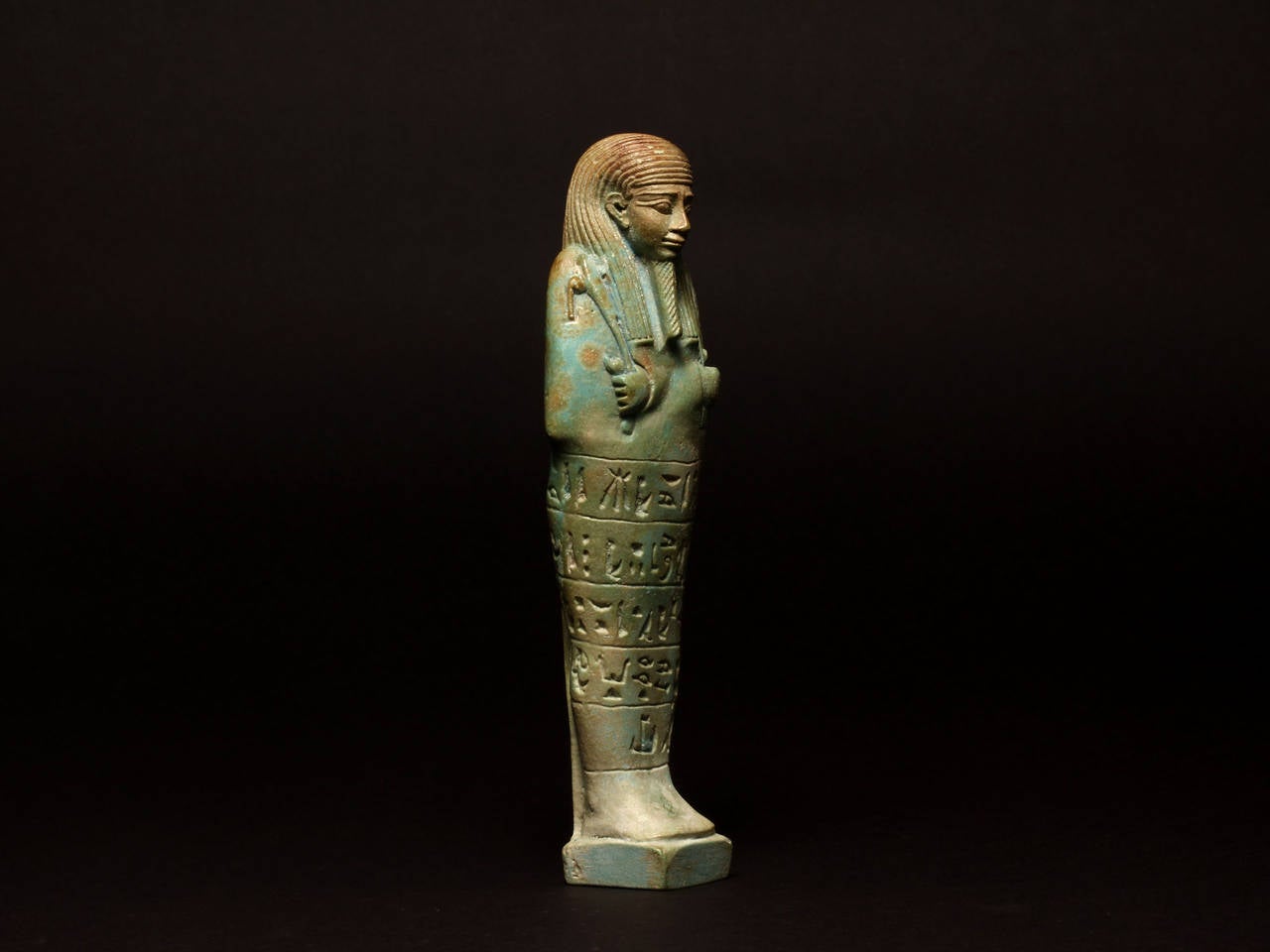 Greenish-blue glazed faience shabti for Horinebesh, standing on an integral raised plinth, decorated with a striated lappet wig and braided beard, the sleeved arms with emerging hands crossed over the chest to hold the pick and hoe, and a parallel