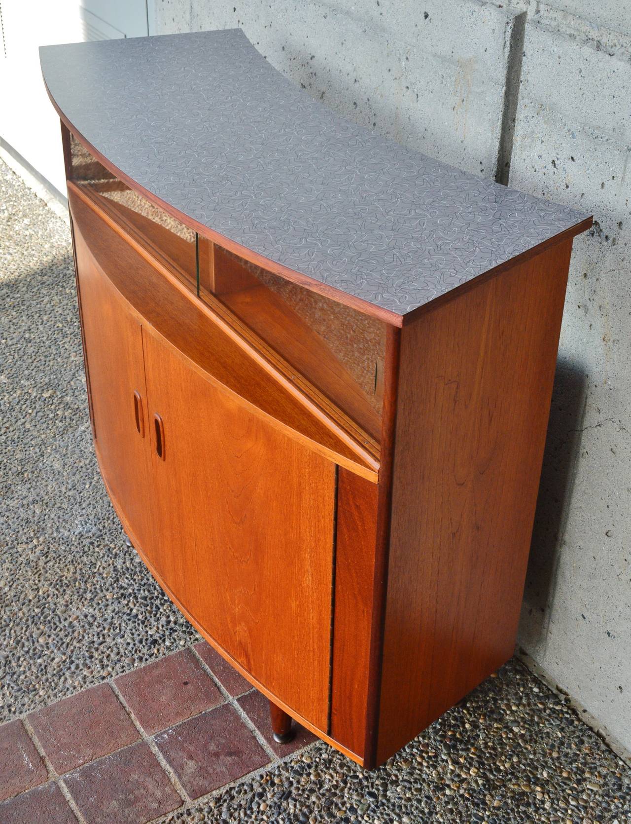 This is a totally cool and unique Danish Modern teak bar! Featuring a bow-front profile, where the front doors and bar top are curved outwards, and the back of the bar top curves inwards. The top was damaged formica, so we replaced it with new