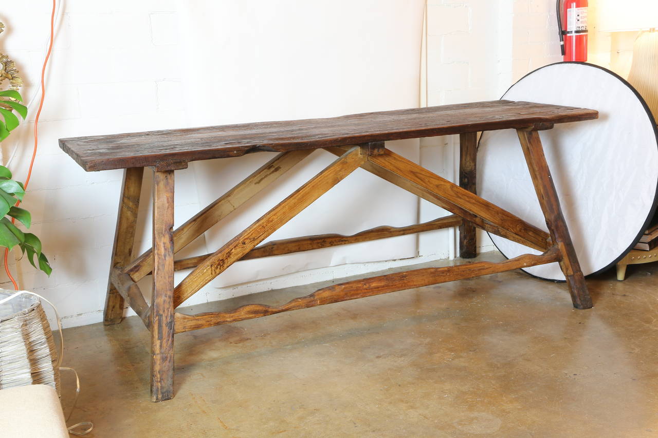 Rustic hand-hewn country farm or work table, 19th century, a plank top raised on a stretcher joined angular supports, the top with holes, heavy use, mortise, tenon and peg joinery.