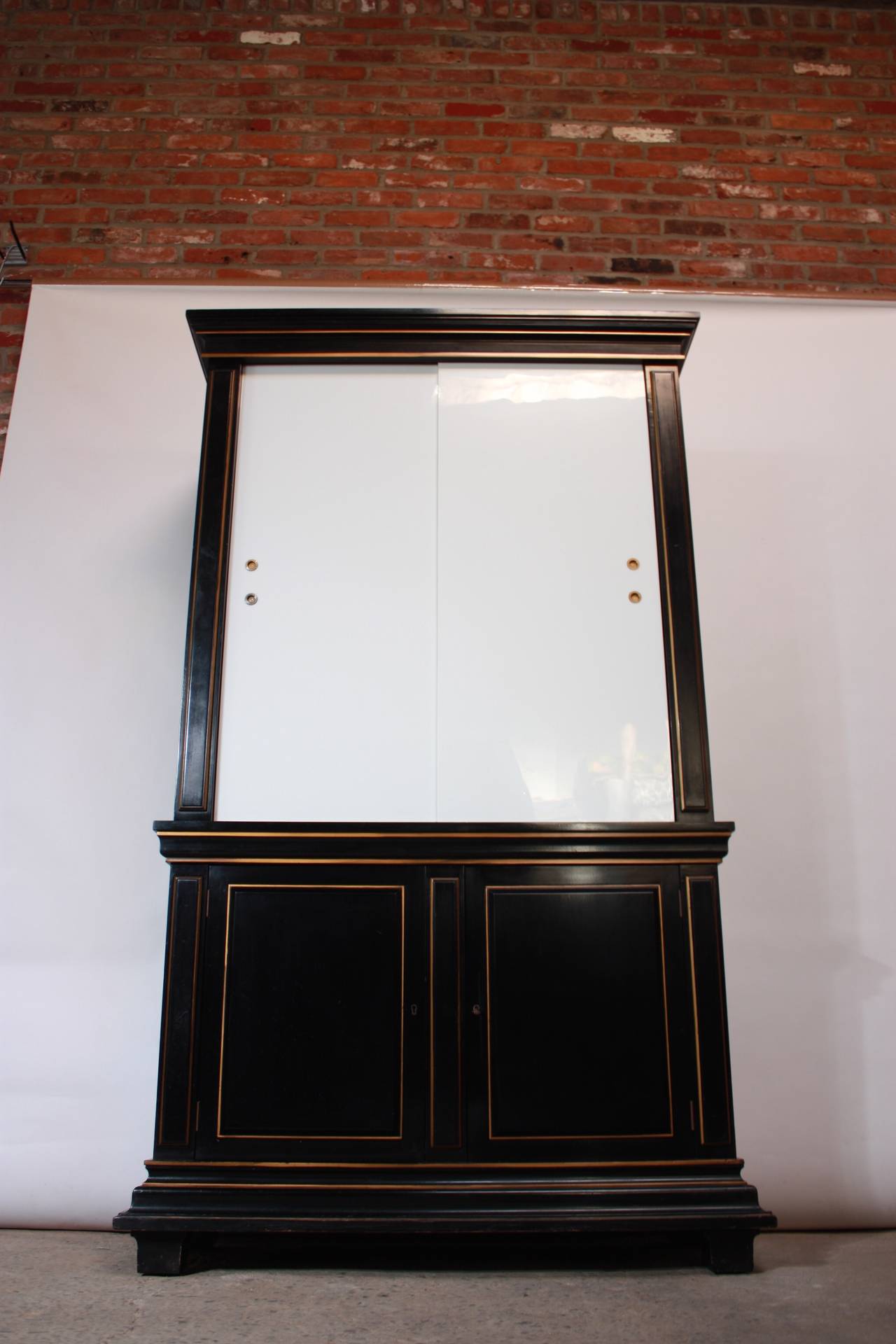 This majestic armoire in the style of James Mont features two sliding milk glass doors, a black lacquered frame, three adjustable shelves on the top, and one built-in shelf on the bottom. The two bottom doors open to reveal the one built-in shelf,