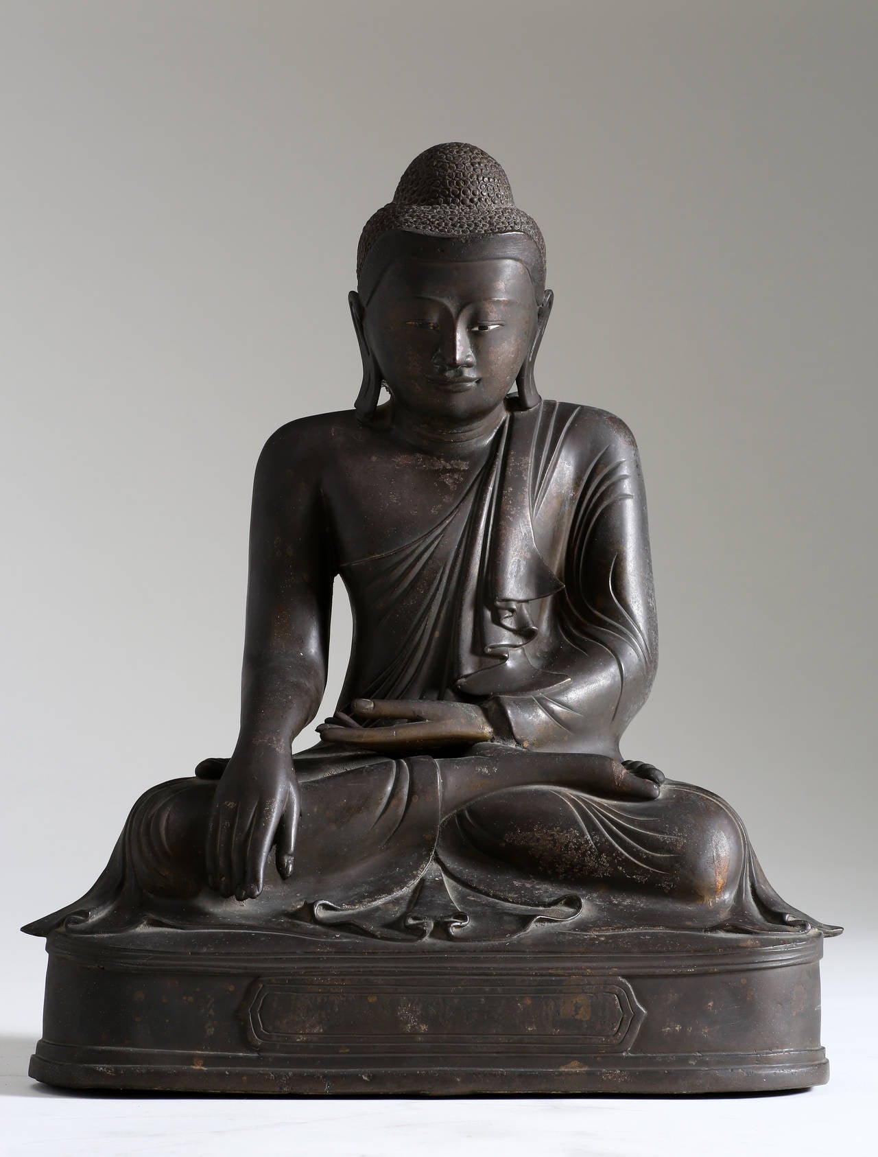 The Buddha is seated on a low base in vajrasana with bhumisparshamudra, in which the finger-tips of the right hand touch the earth to symbolize the moment of enlightenment. The Buddha is clothed in simple monk’s robe with billowing folds of fabric.