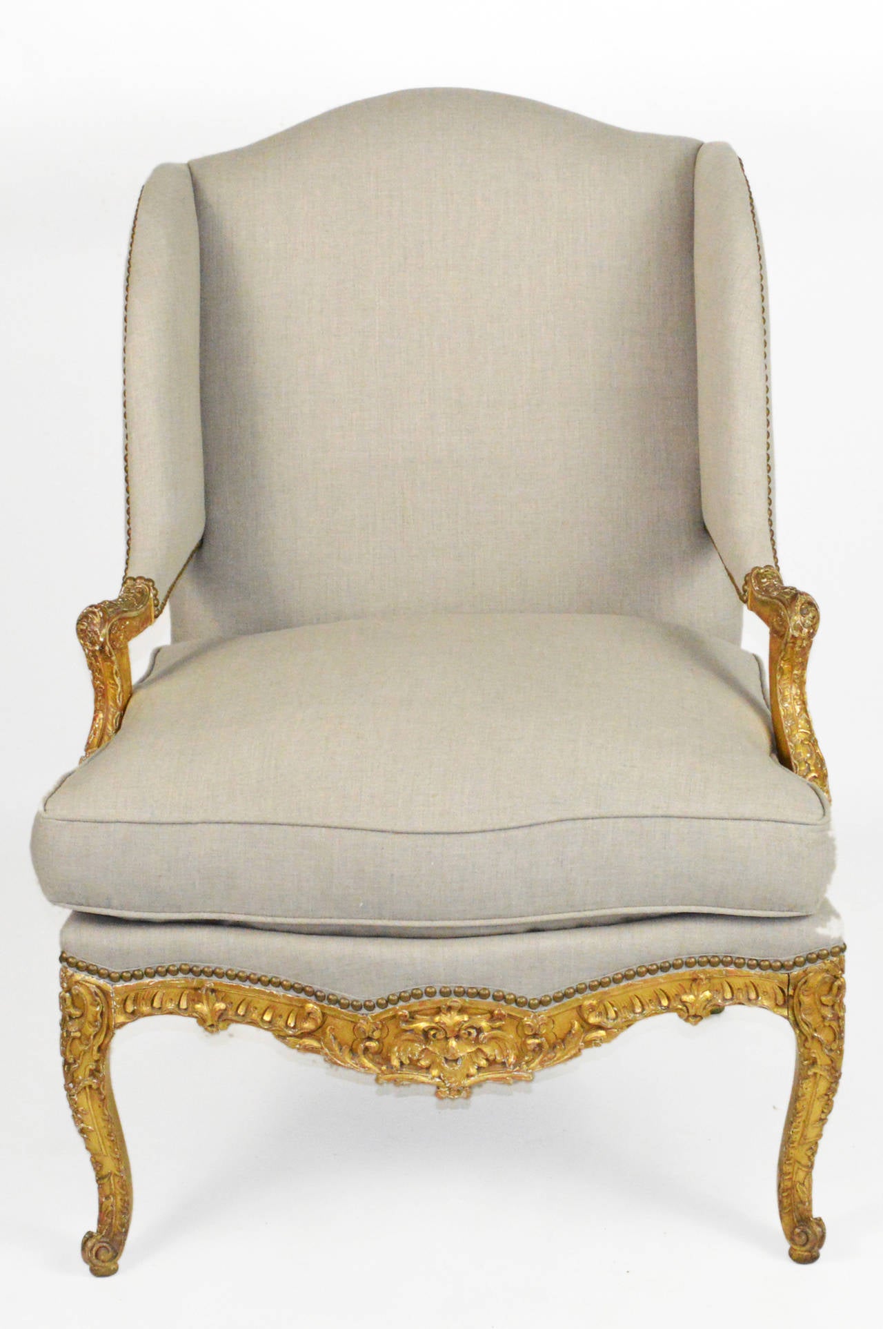 A fine Louis XV style wing chair. Raised on cabriole legs. An intricately carved giltwood frame. Having padded manchettes, a loose cushion, covered in linen with nailhead trim.