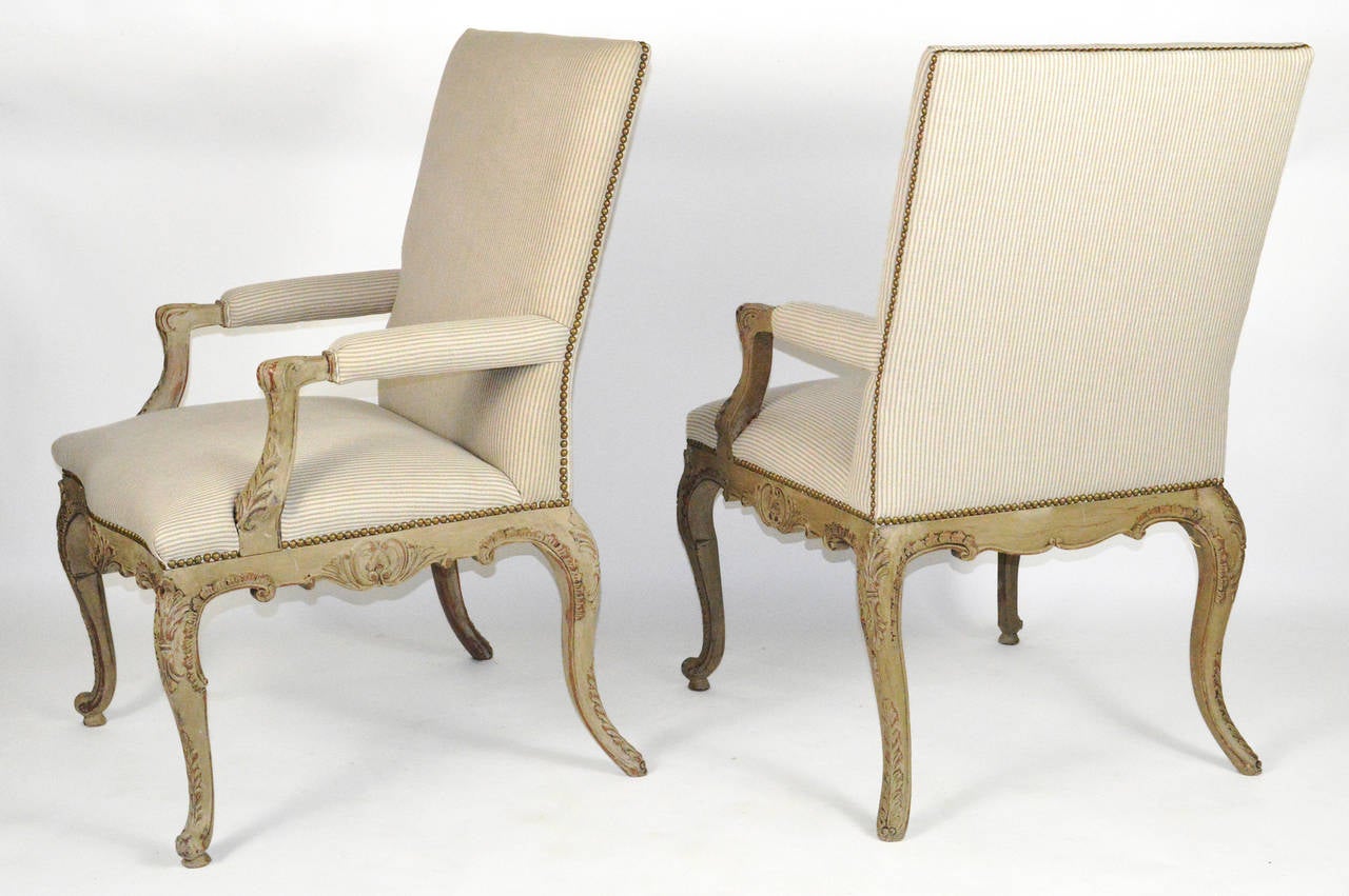 A beautiful pair of French Regence style painted open armchairs. Upholstered arm pads extending to scrolled arm supports, standing on cabriole legs front and back. Upholstered in a striped fabric with nailhead trim.