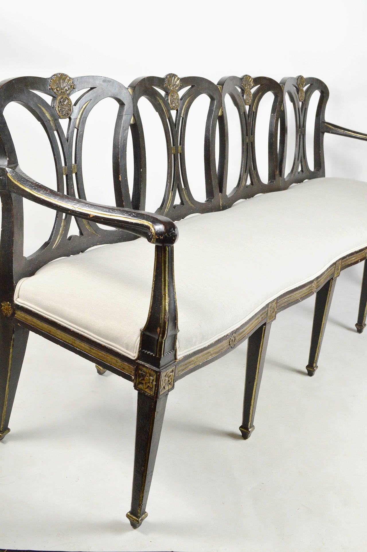 A fine 19th century neoclassical style black painted and parcel-gilt bench. Frame with shell and circular medallion accents on an arched slat back. Bench with serpentine front with gilt accents on apron, above ten tapered legs with spade feet.