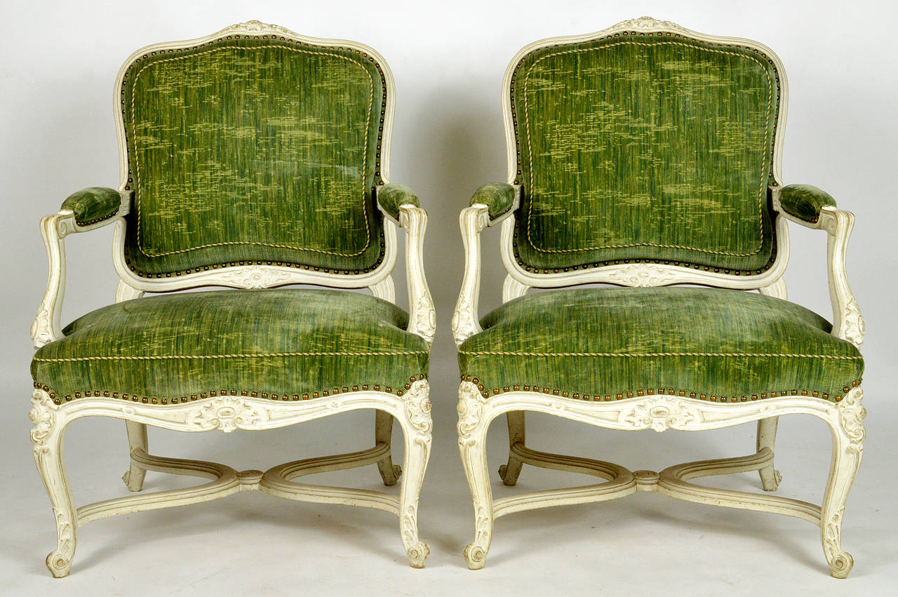 A very fine pair of Louis XV style carved and white painted upholstered chairs. Each having foliate carved crest rails over the upholstered backs, arms and seats, raised on cabriole legs, joined by X-form stretchers.
