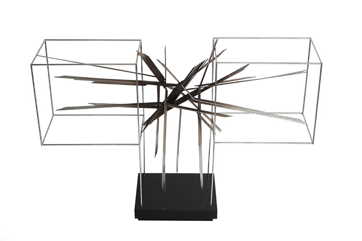 This sculpture features a chrome plated steel wire geometric framework with an abstract Brutalist metal burst suspended within the frame.  The burst is chrome steel with darkened areas.  Black painted wooden base.  Plaque on base with signature has