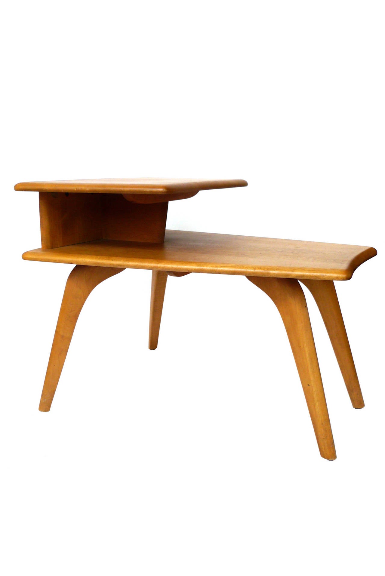 This Midcentury end table is designed by Heywood-Wakefield. Crafted from ashwood, the table has a beautiful wheat finish. Its sculptural form includes two flattops, the upper tier being smaller than the bottom. The legs curve at the top and taper