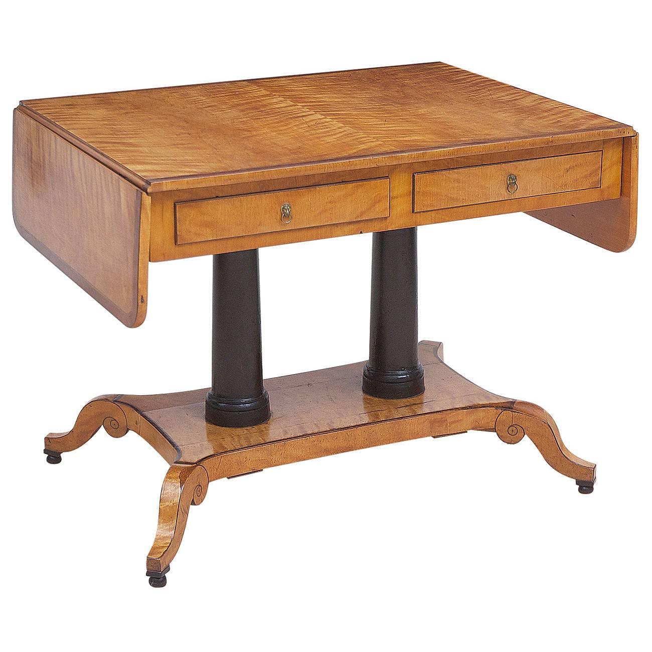A sofa table or writing table in birch with mahogany bandings and ebonized columns resting on base terminating in carved 