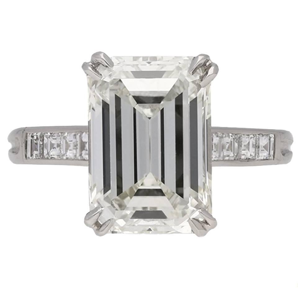 Emerald-cut diamond ring. Set to centre with an octagonal emerald-cut diamond, K colour, VVS2 clarity, in an open back double claw setting with a weight of 4.54 carats, further enhanced by eight square step cut diamonds in open back rubover channel