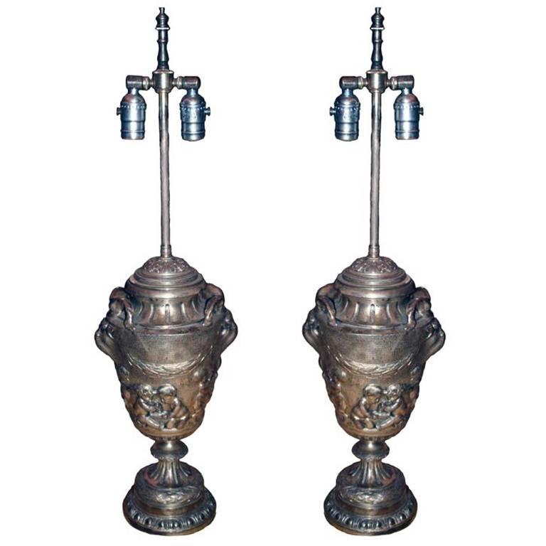 A pair of French bronze urns fitted as lamps, in the manner of Colodion (1738-1814) the ovoid form body chased with scenes with classical maidens,  flanked by roaring animal mask handles, the spreading socle with stiff leaf cast surround resting on