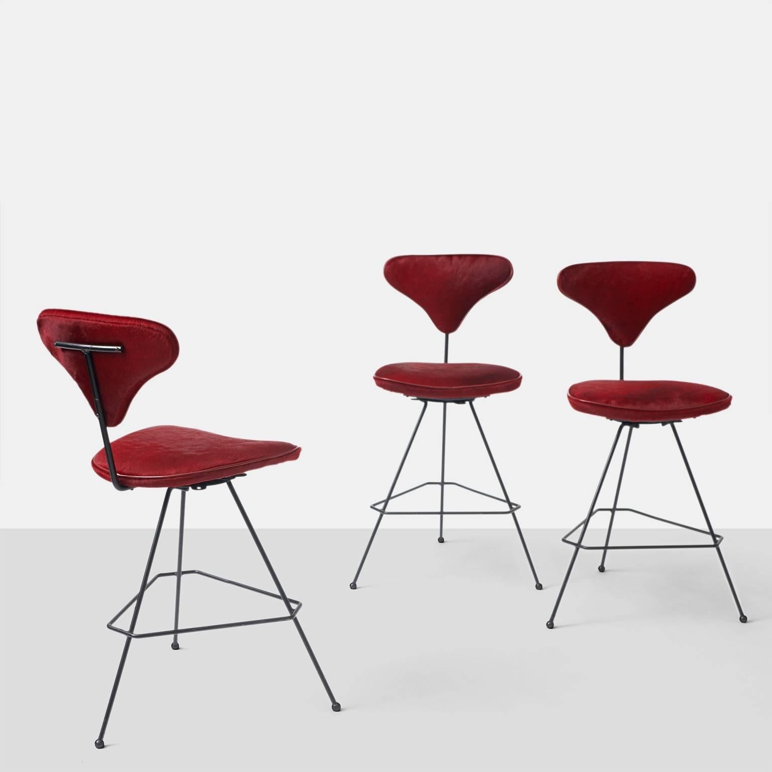 A group of three bar stools with a swivel base and black iron frame. The seat and back have been upholstered in a red hair on hide with red leather trim. The stools were made for Elton in 1950.
