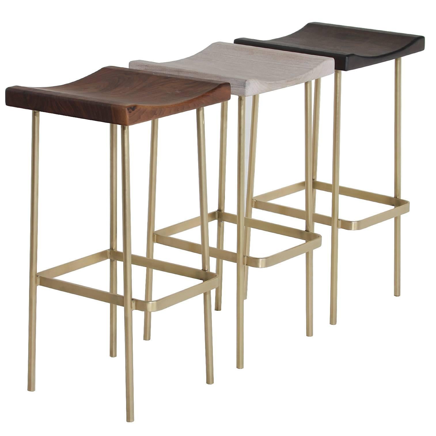 The Bundinha stool by Thomas Hayes Studio with brass frame, with a variety of wood tops and a curved flat bar footrest. A smaller version of our Bunda stool with a comfortable contoured square seat. Finishes from left to right: Pure oil bleached,