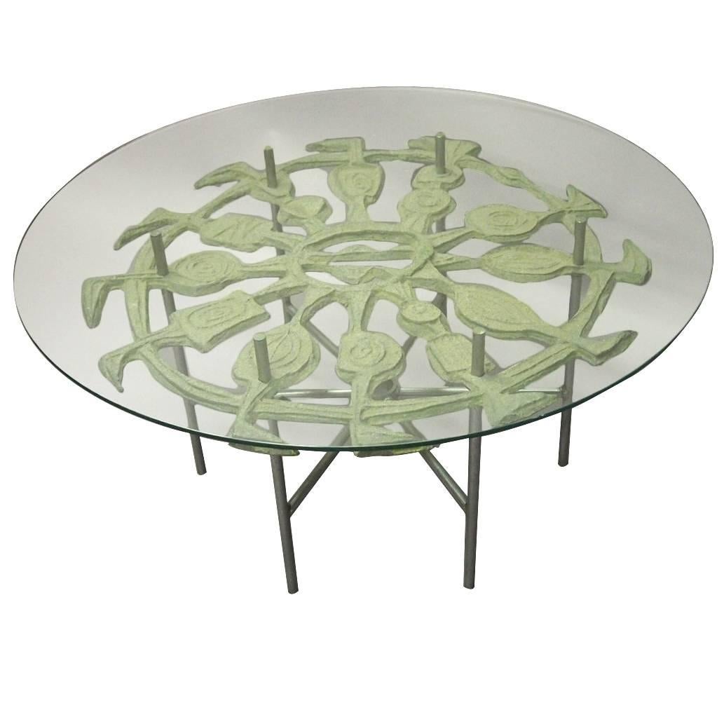 Coffee table has a tubular metal base that supports a decorative cast aluminium design with a grey paint finish below a round glass top.