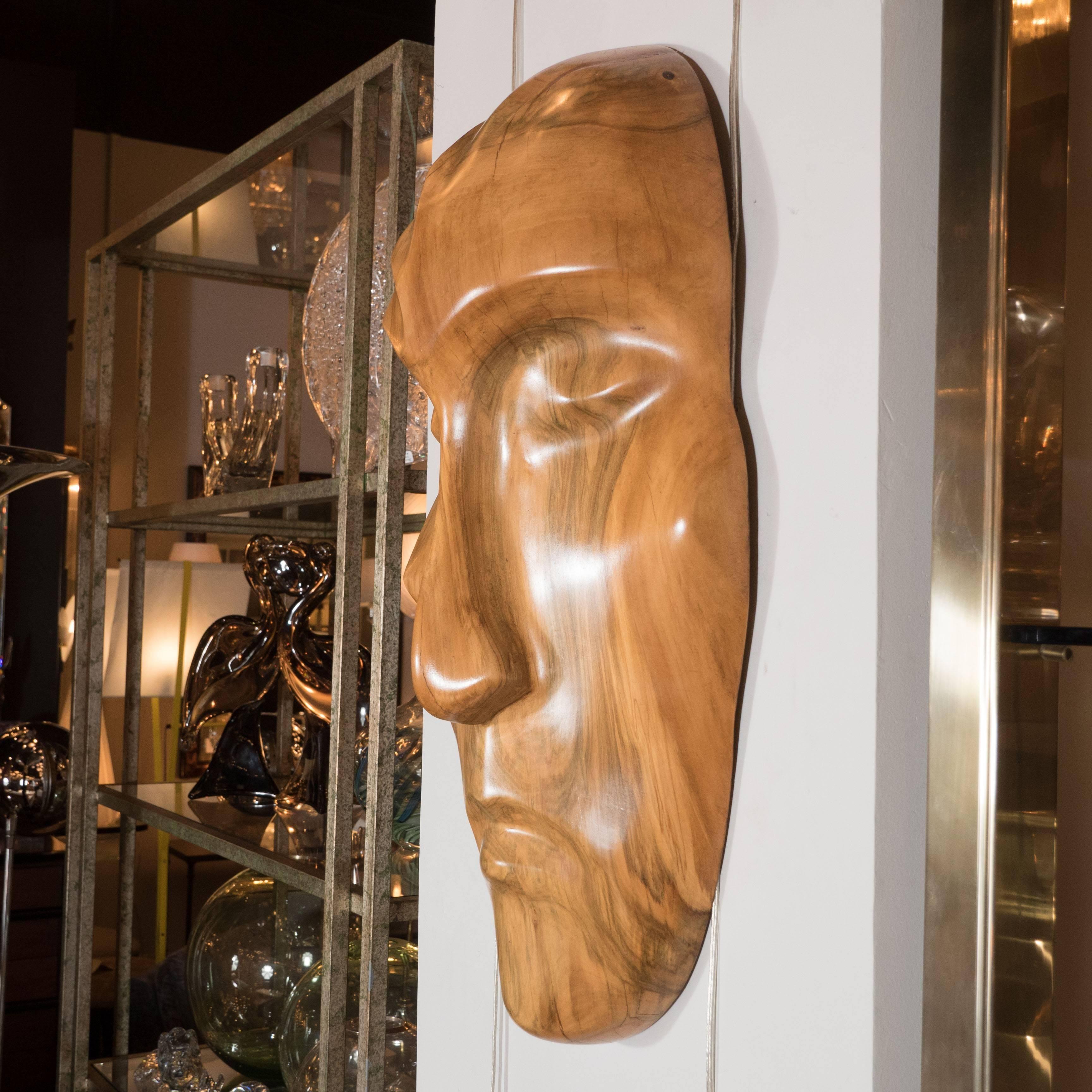 This striking Mid-Century Modernist mask in hand carved exotic honey hued wood reads as both tribal and contemporary. The face- reminiscent of a Haida mask- exhibits a serene expression, evocative of a sleeping state. This sculpture embodies the