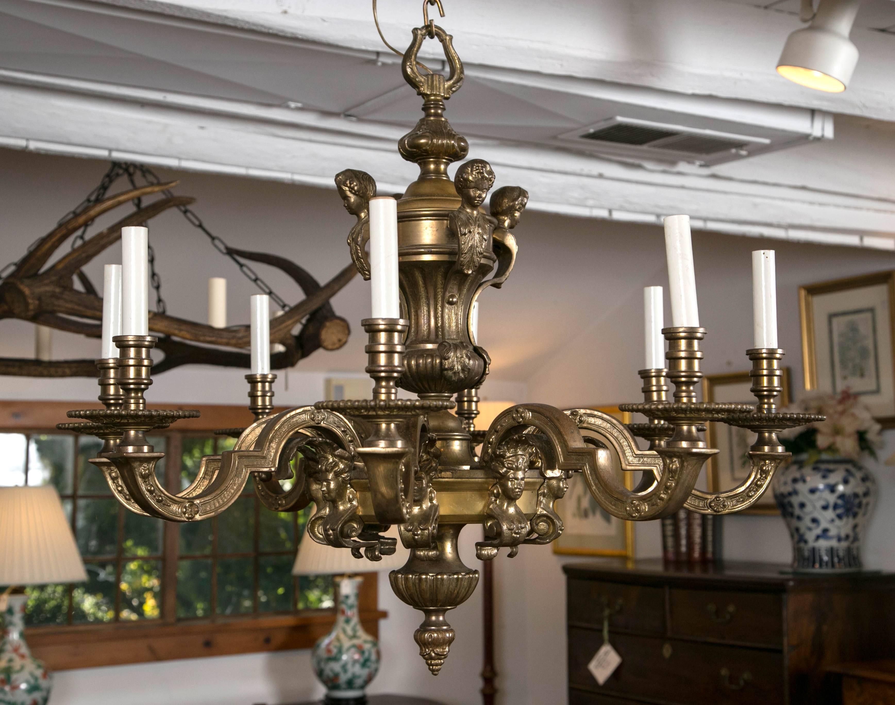 Festooned with busts above and below its bold, architectural arms, this chandelier puts the classic in neoclassic. The eight arms radiating from a central disk and the urn post above leads the eye up to a magnificent lyre-shaped connecting hook.