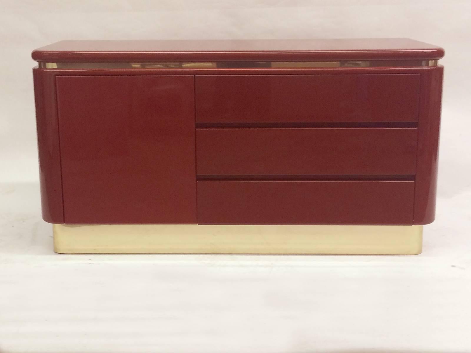 This is a deep red lacquer Hollywood Regency dresser.  The dresser has three drawers and one cabinet door . Is is wrapped below the top and on the base in brass.   
