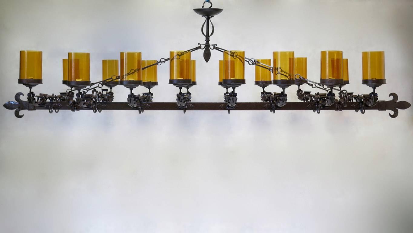 Très impressionnant lustre à seize lumières en fer forgé de style médiéval, de très grandes proportions, se terminant par des fleurs de lys stylisées. Chaque lampe est dotée d'un grand abat-jour en verre ambré. 
Il s'agit d'une pièce rare.