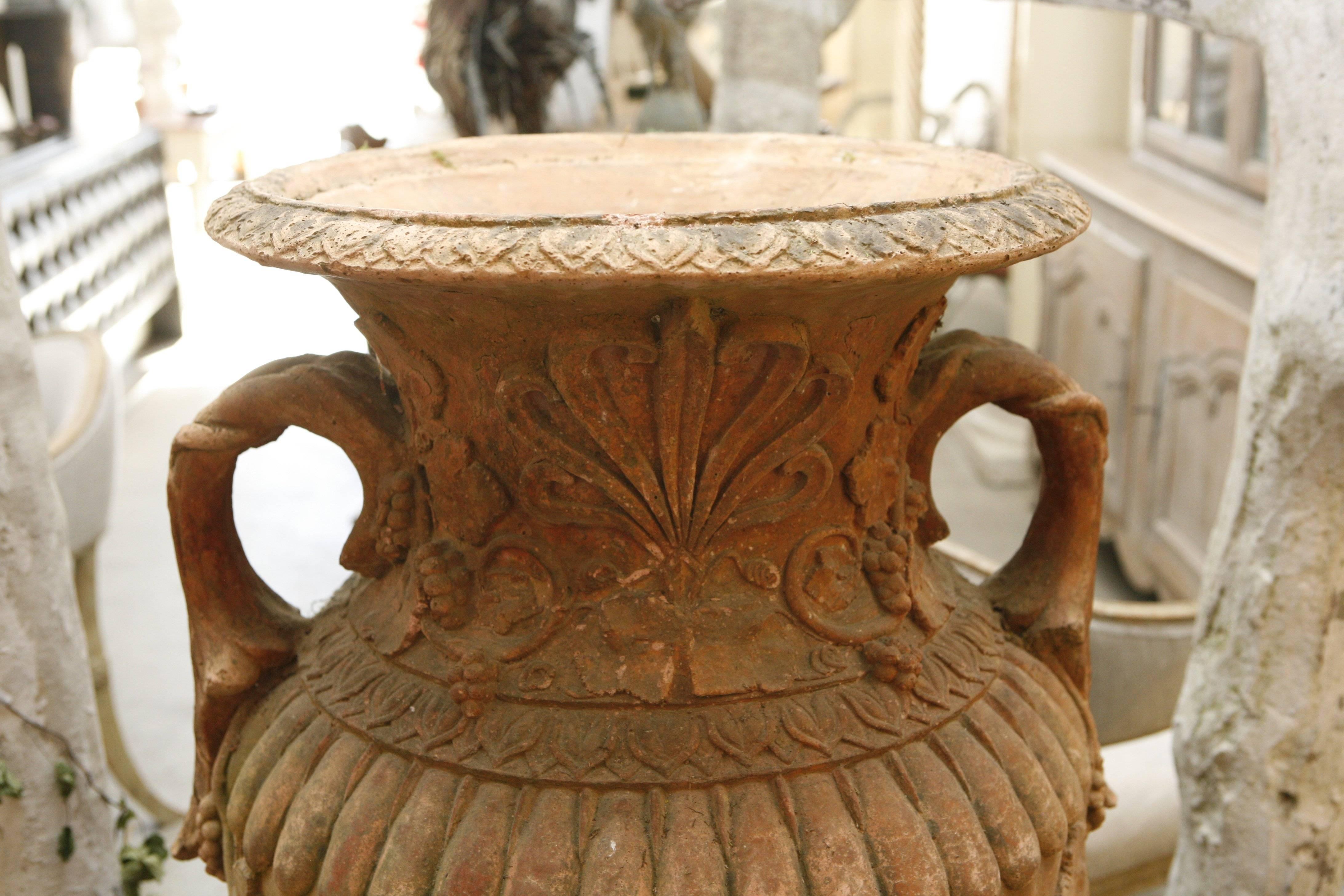 A single large red terra cotta urn with floral decoration sitting on a pedestal. Exposure to the elements from being outdoors has caused beautiful patina.