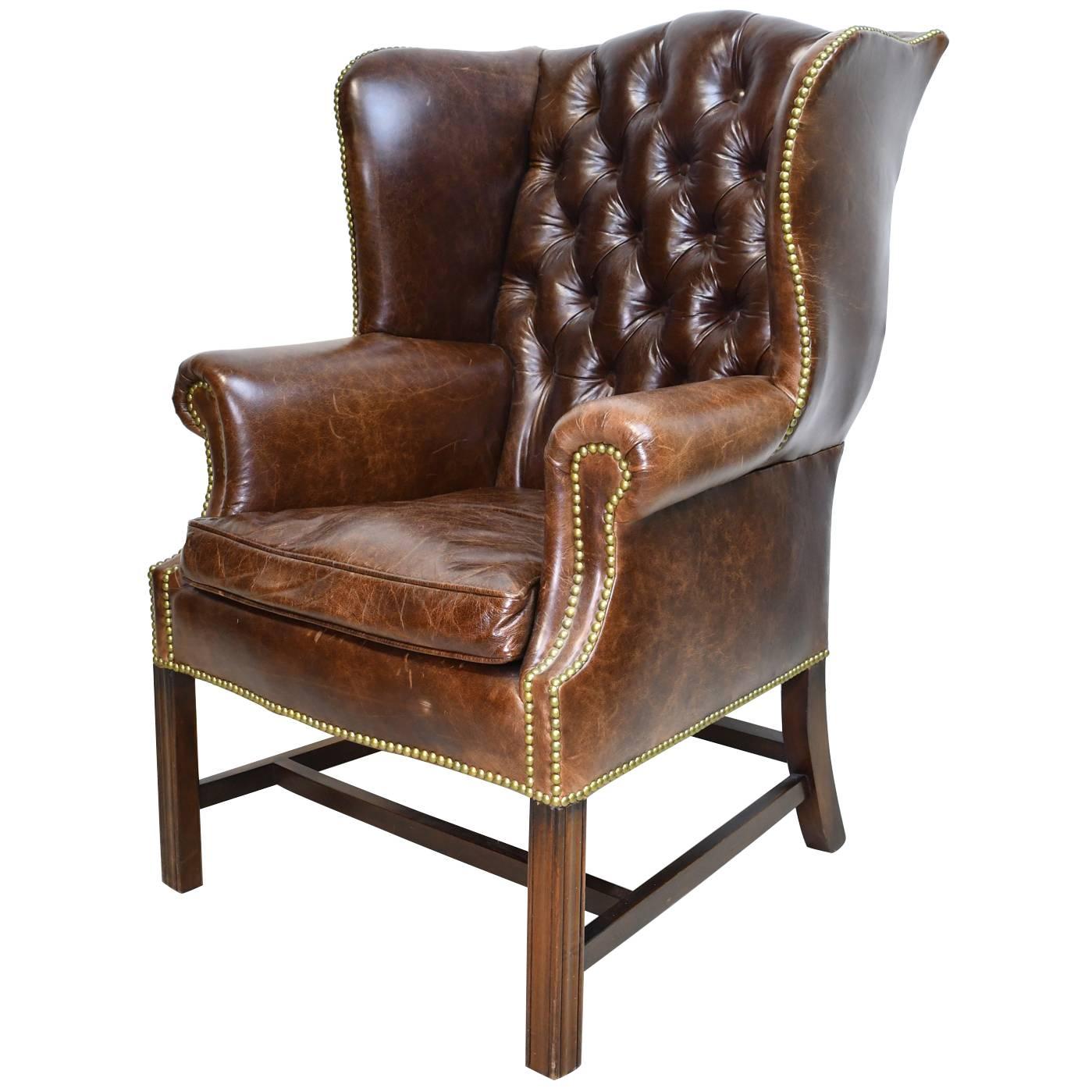A classic English-style Chesterfield wing-back chair upholstered in a medium brown leather (milk chocolate color) with tufted back & brass decorative nail heads around the silhouette. Chair rests on square Marlboro front legs & splayed back legs