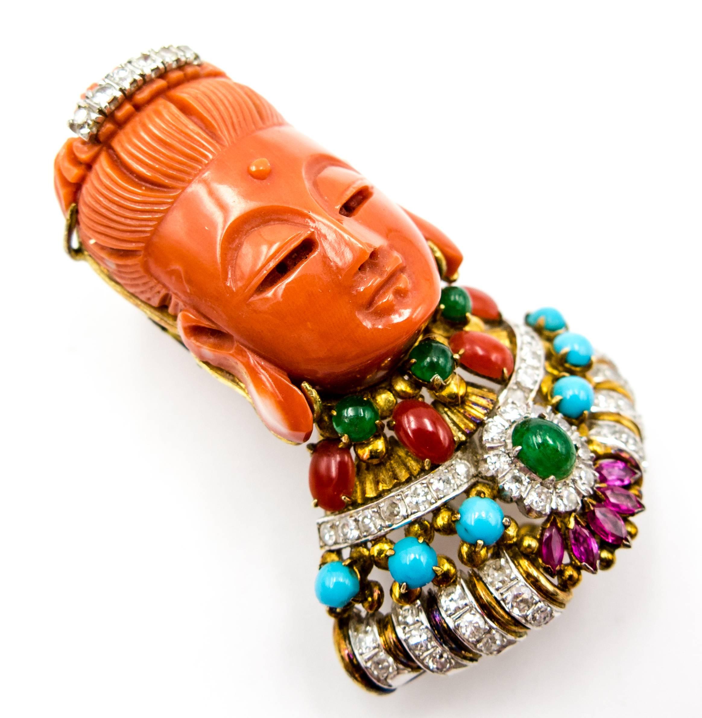 Serene and impassive, this lovely head made of carved orange coral is accented at the top by a diamond and white gold headdress and at the bottom by a cape made of yellow and white gold set with oxblood coral, turquoise, emerald, diamond, and ruby. 