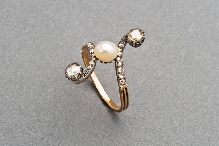 A French scrolling diamond and pearl ring of rococo design set with an oval pearl, the curved shoulders enhanced with lines of rose diamonds, each ending in an oval rose diamond, mounted in silver and 18k rose gold.

Circa 1890, unmarked except for