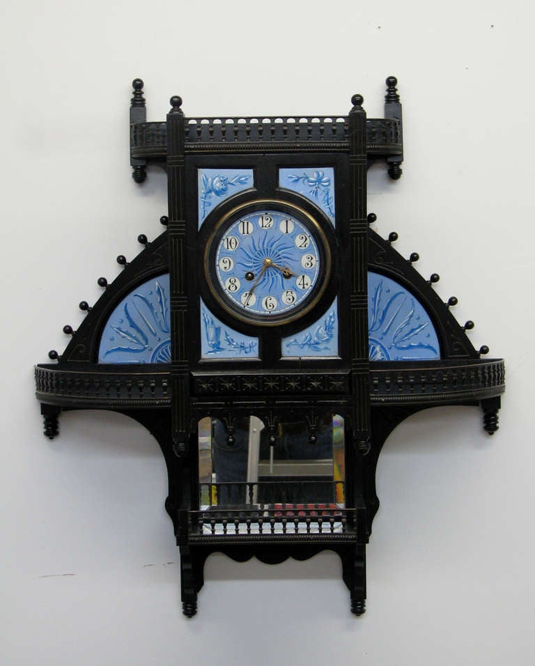 A very unusual Aesthetic Movement ebonized wood wall clock having openwork balustraded shelves backed by beveled mirror and decorated chalcedony blue ground ceramic plaques; also having a similarly colored and decorated ceramic dial the design of