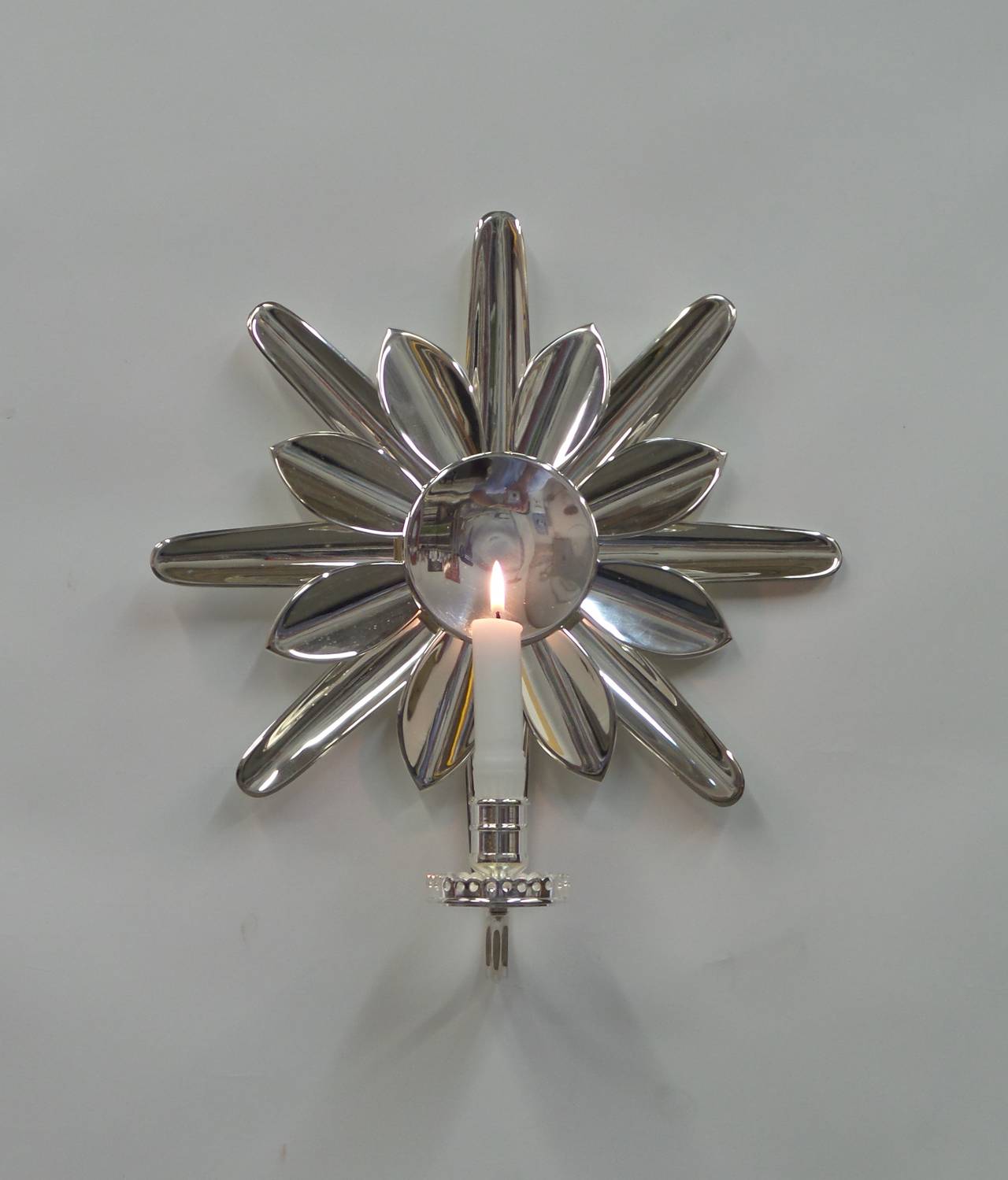 A sunburst form wall sconce, in silvered brass, with a single candleholder arm. Wiring with a wax faux-candle stick and candle form bulb available.