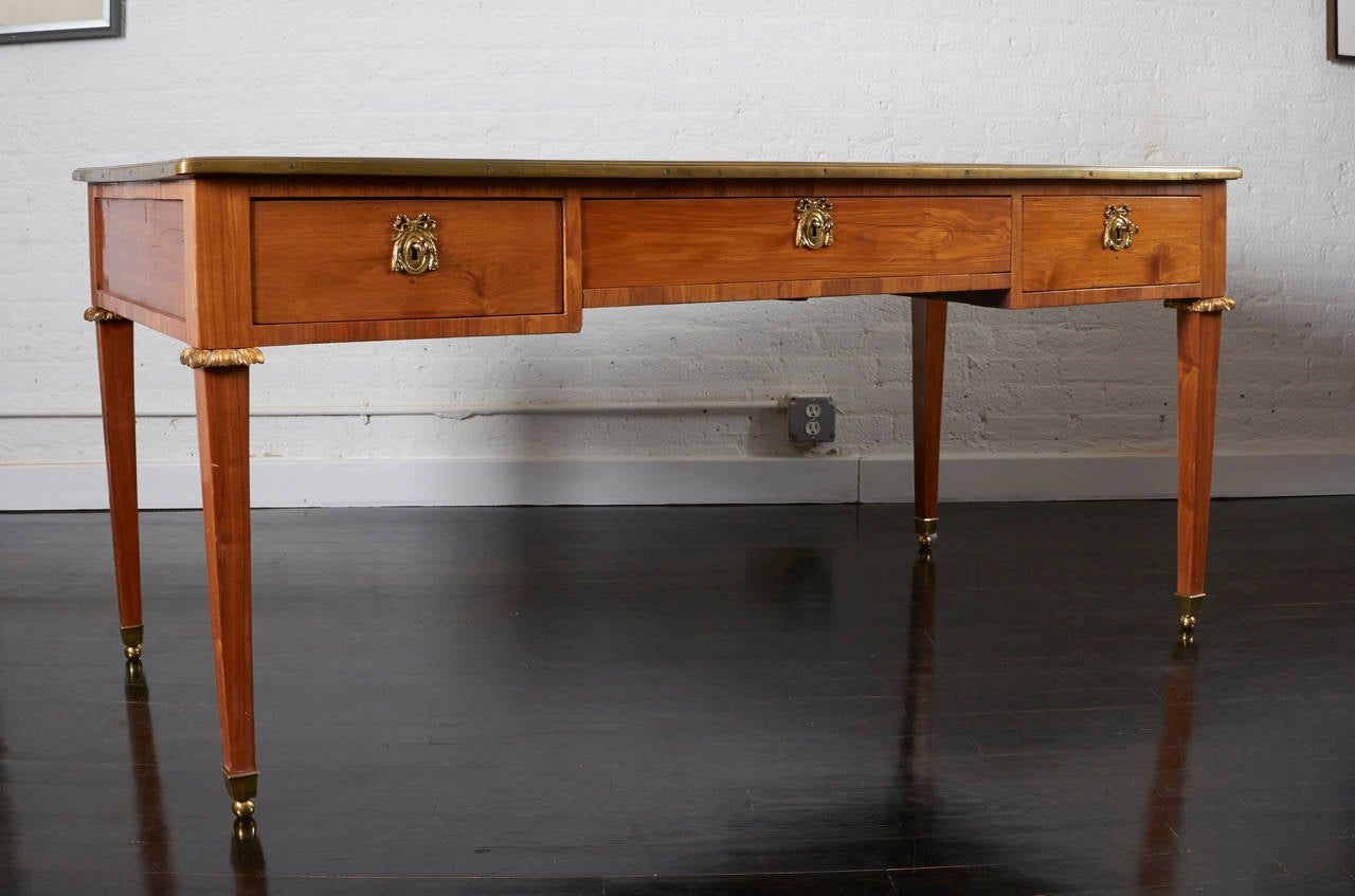 An 18th century French bureau plat made of beautiful fruitwood with brass details and a distressed leather top edged on a brass border all around.
There are three drawers with ormolu mounts around a working lock, the legs begin with a brass palm