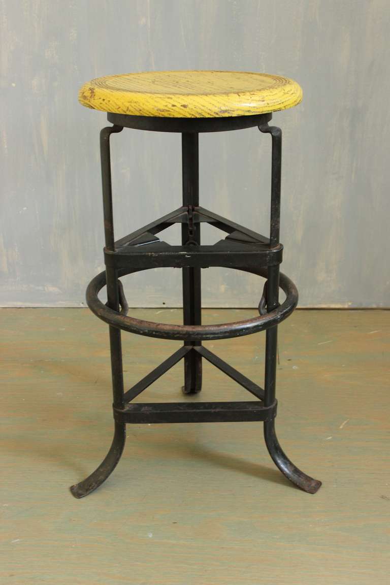 An American early 20th century adjustable black iron industrial factory stool with a  yellow wooden seat. Add unique character to any room with this black iron industrial factory stool. Featuring a striking yellow wooden seat, this stool is full of