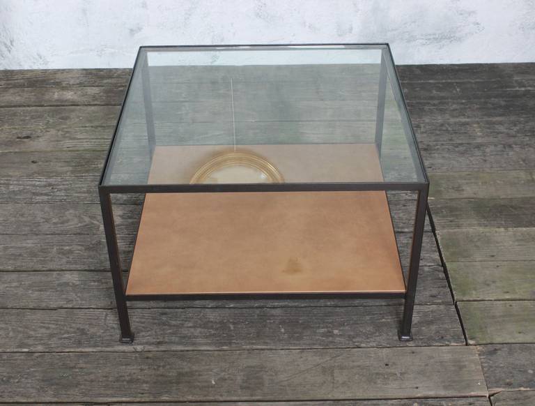 Square iron frame coffee table in bronze paint finish. Top shelf is clear glass and the bottom shelf is distressed leather wrapped wood. This is a floor sample from our discontinued Reeditions line. The sample is in good condition and is ready to be