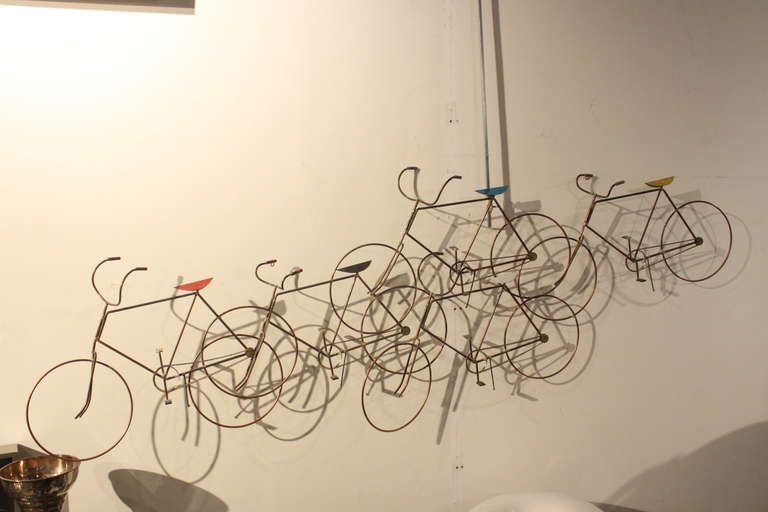 This great decorative wall sculpture by Curtis Jere depicts a group of bicylces fabricated from brass with primary color and black and white seats. Shown hanging from a hook, this wall sculpture hangs well balanced from two eye hooks