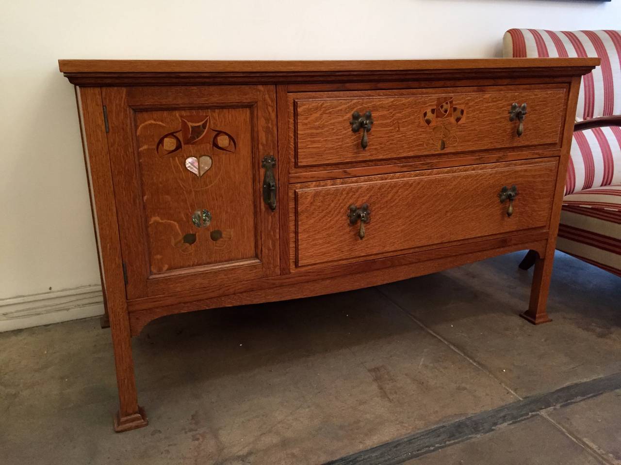 A beautiful Arts and Crafts cabinet done in the Glasgow style. It is done in golden oak with numerous mother-of-pearl and wood inlays and contains two drawers and two shelves. Tear drop brass pulls. Maker's mark. Matching pieces available.